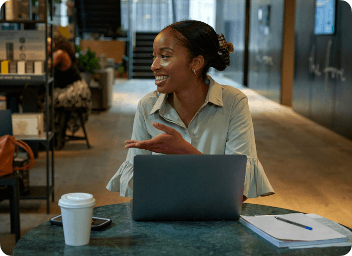 Kvinna ler och gestikulerar engagerat vid ett bord med en kaffemugg, anteckningsblock och laptop i en öppen kontorsmiljö.